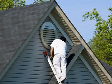 Man on ladder repairing or inspecting a house vent under the roof.