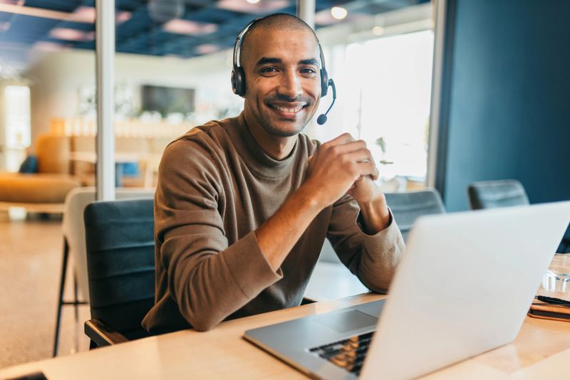 Businessman wearing headset and clasping hands on work desk with laptop, smiling confidently at camera in call center office