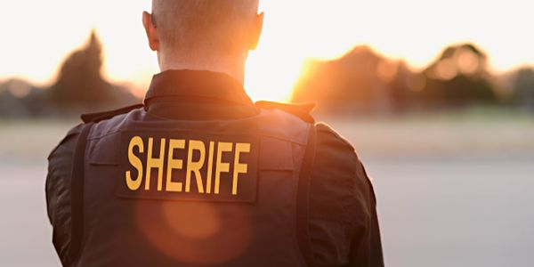 Sheriff in tactical vest facing a sunset outdoors.