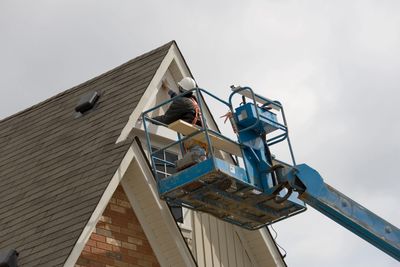 Worker painting house trim from a blue lift on a cloudy day.
