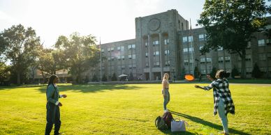 People playing frisbee on a grassy field near a large building.