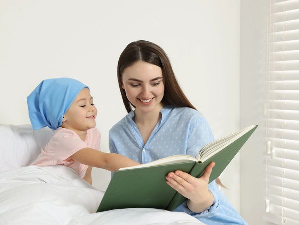A woman reads a book to a child wearing a blue headscarf in bed.