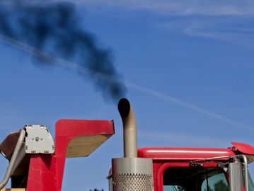 Black smoke emitting from a red truck's exhaust pipe against a clear sky.