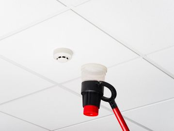 A tool with a red handle is used to reach a smoke detector on a white ceiling.
