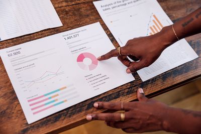 Person pointing at charts and graphs on printed data sheets on a wooden table.
