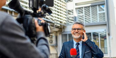 A smiling male reporter with a microphone being filmed outdoors.