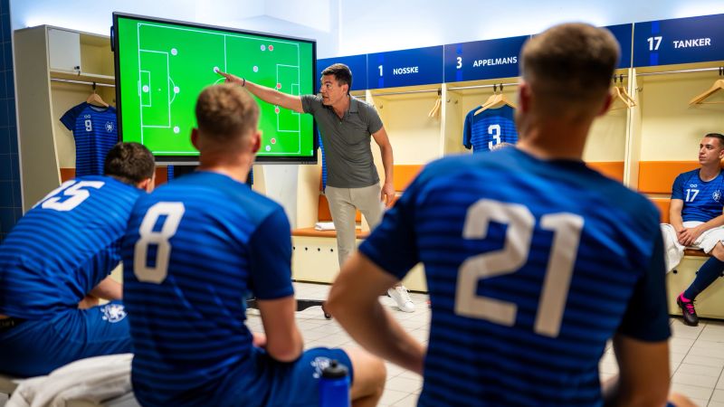 Male soccer coach showing players positioning on large interactive screen, planning game strategy in locker room