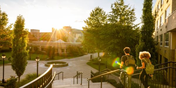 Two young people walk down steps in a sunlit campus courtyard.