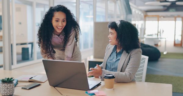 Two women collaborating and discussing work at a laptop in a modern office.