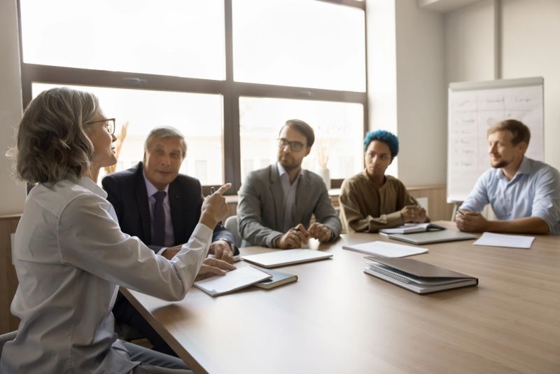 Diverse business team listening to mature grey haired project leader, manager woman, meeting at large table in boardroom, discussing collaboration, sales reports, working together