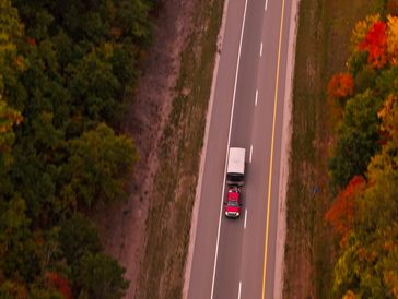 A red truck towing a trailer on a road surrounded by autumn trees.