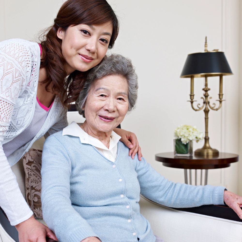 A younger woman embraces an elderly woman seated on a couch, both smiling warmly.