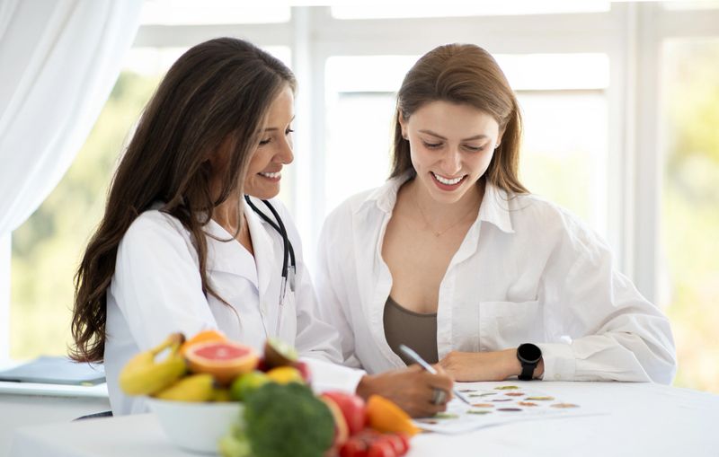 Cheerful adult caucasian nutritionist doctor in white coat makes notes of weight loss plan to young woman patient in clinic interior. Health care, proper nutrition, recommendation by professional