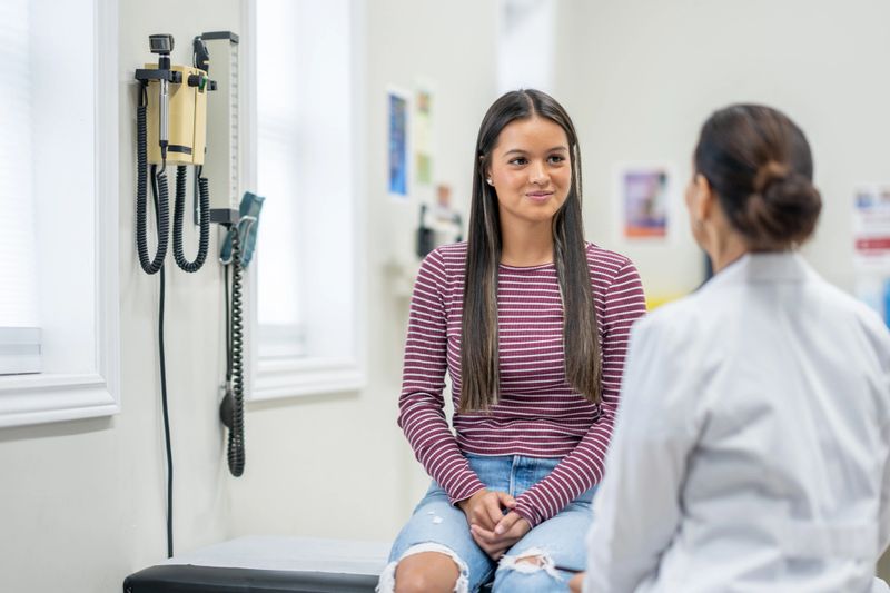 A young female teenager sits up on an exam table during a routine medical appointment with her doctor.  She is dressed casually and talking with her doctor who is standing in front of her.