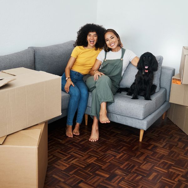 Two women and a black dog sitting on a gray couch surrounded by moving boxes.