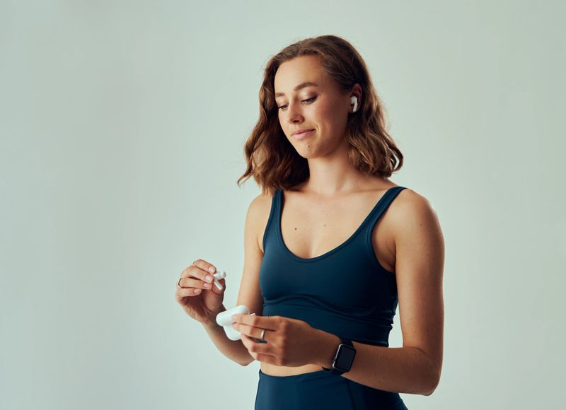 Young caucasian woman with fitness tracker looking down while holding wireless in-ear headphones
