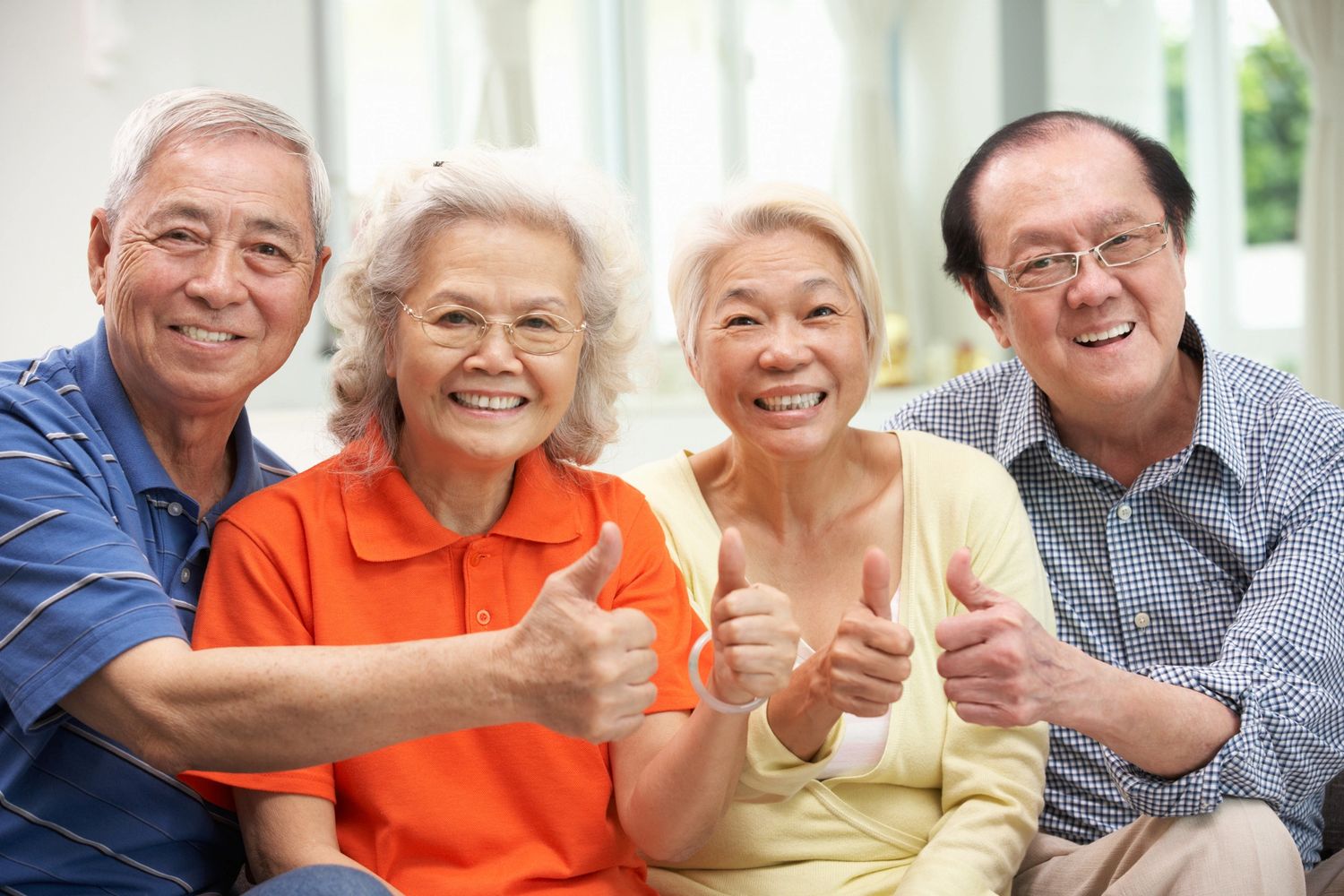 Four elderly friends smiling and giving thumbs up in a bright room.