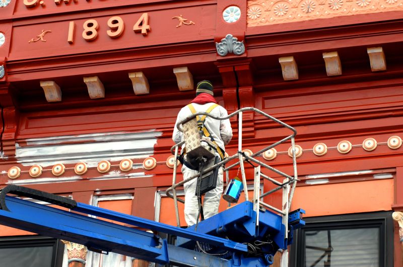 Painter applies new paint to historic building in Calumet, Michigan.  He is standing on a blue lift and building has date of 1894.