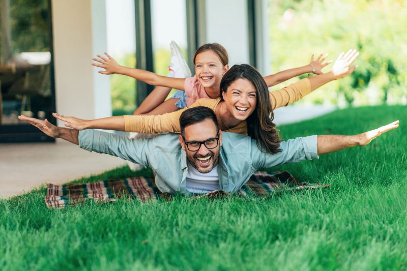 Fun portrait of smiling father, mother and daughter  laying on the grass in front of their house. Family stuck on top of each other with arms stretched out pretending to be a plane. Playing outdoor is the best bonding.