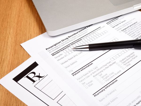 Pen on prescription drug reimbursement form next to laptop on wooden table.