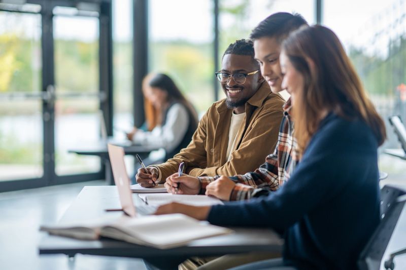 Three University students are seen sitting together in class as they work together on an assignment.  They have a laptop open between them as they each give input for the assignment.