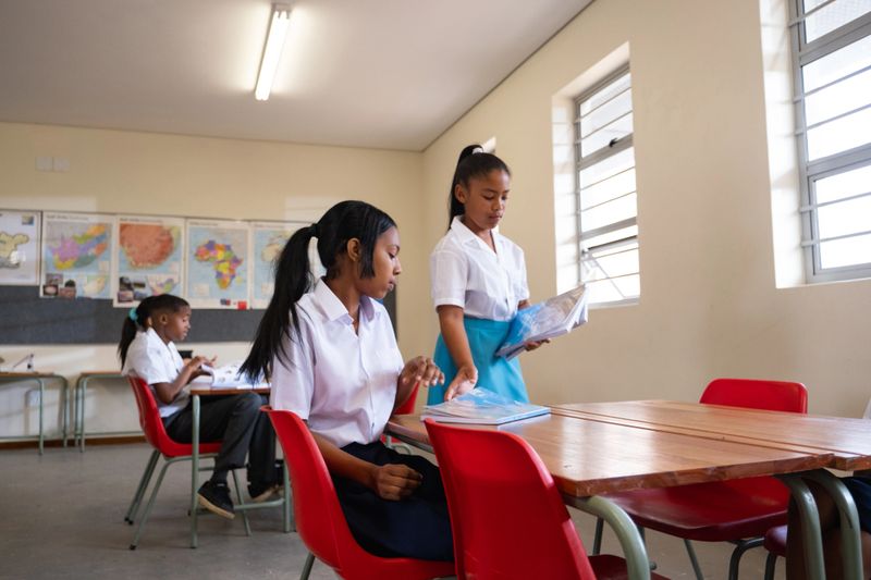 A young African schoolgirl helps her teacher by handing out textbooks to her fellow classmates.