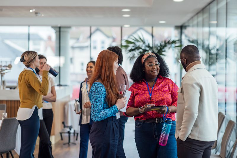 A wide angle view of a group of mixed age professionals who are taking advantage of a networking opportunity at a business conference in the North East of England. They are stood in smaller groups and getting to know each other during the networking getting to know you breaks of the conference. Some are carrying their water bottles and the others their notebooks and they are wearing their ID lanyards.