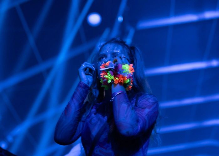 Large concert venue filled with a crowd of people. Performers on stage and audience cheering. Strong neon lighting and smoke effects
