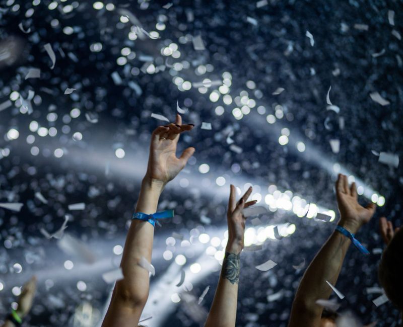 Large concert venue filled with a crowd of people. Performers on stage and audience cheering. Strong neon lighting and smoke effects