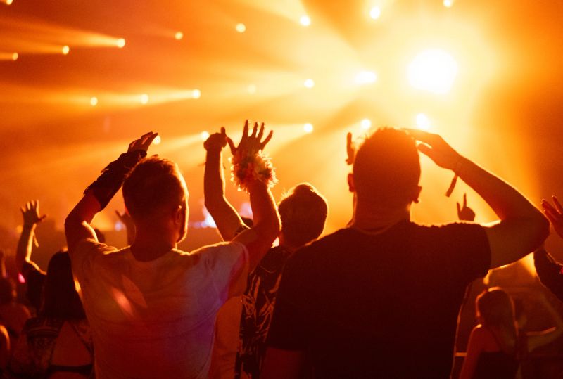Large concert venue filled with a crowd of people. Performers on stage and audience cheering. Strong neon lighting and smoke effects