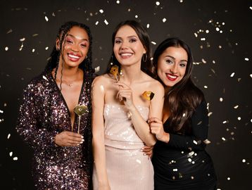 Three women in glamorous outfits celebrating with confetti and gold props.