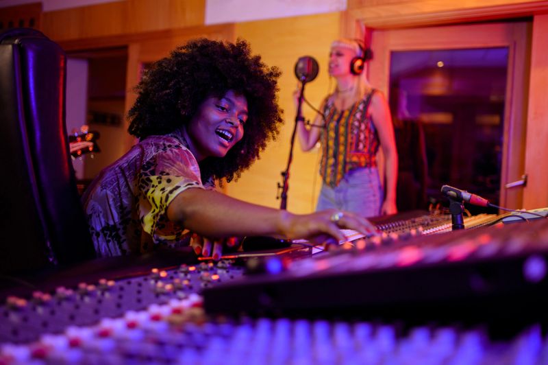 Portrait of a smiling multiracial female music producer operating her music console while female vocalist is being recorded. A happy sound technician is sitting in a recording studio and recording.