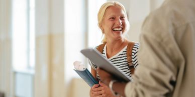 Two students happily chatting in a bright hallway with books and a laptop.