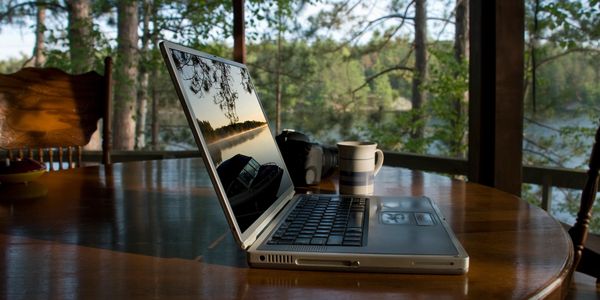 Laptop on wooden table with scenic lake view reflected on screen.