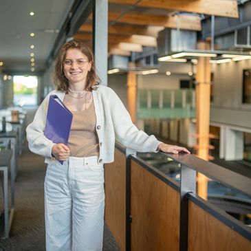 Young woman with glasses holding a purple folder in a modern indoor space.