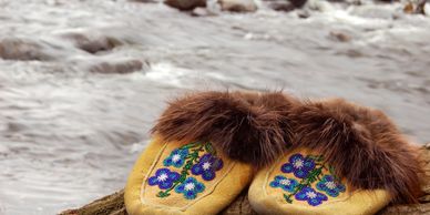 Traditional fur-lined moccasins with blue floral beadwork on a log near a river.