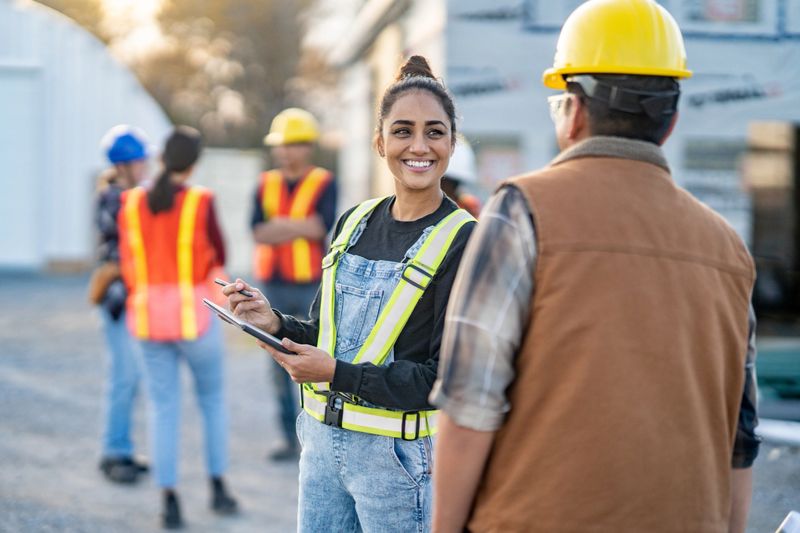 A female foreman reviews plans electronically on a tablet with a crew member as they review the site tasks together.  Other crew members can be seen conversing in the background.