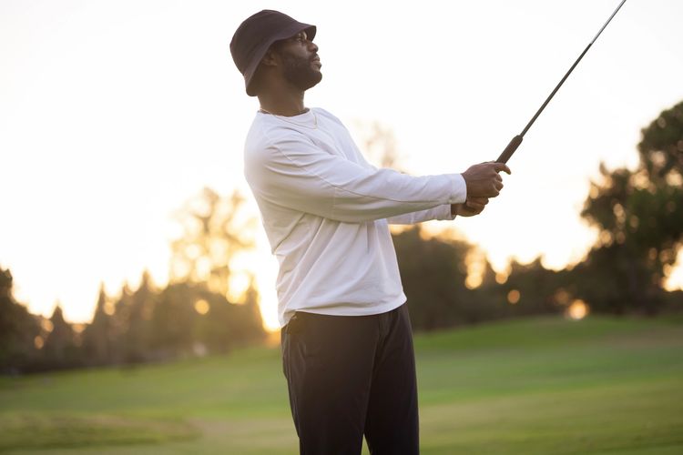 Man in bucket hat swinging a golf club on a course at sunset.