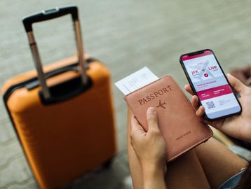 Traveler holding a passport and mobile boarding pass beside an orange suitcase.