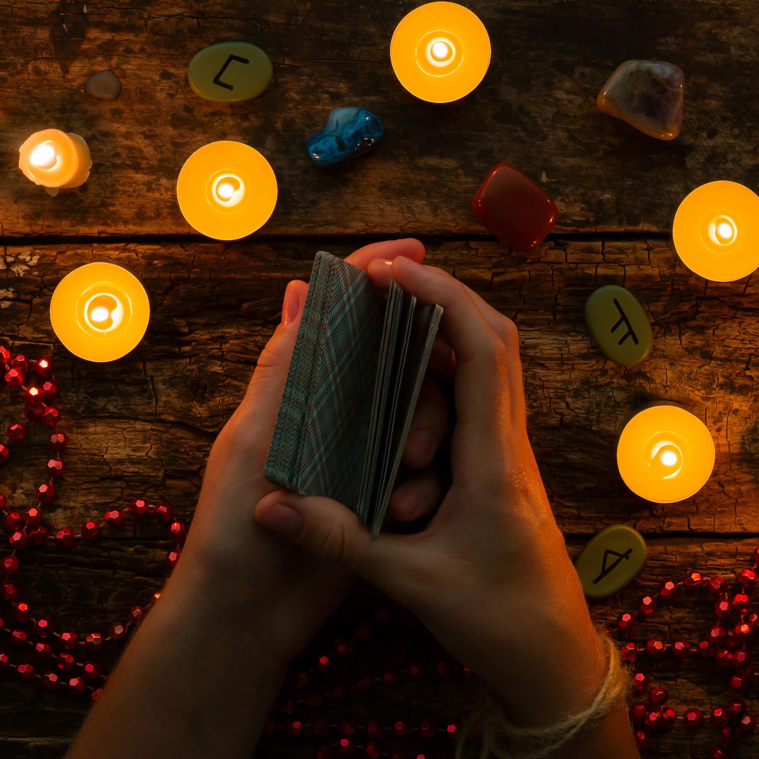 Hands shuffling tarot cards surrounded by candles and rune stones on a wooden table.