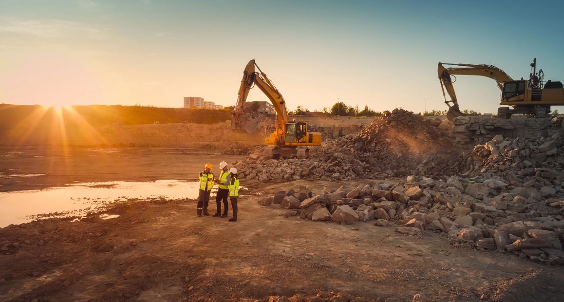 Construction workers discuss at a rocky excavation site during sunset.