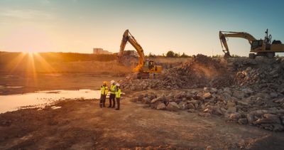 Three workers in safety gear discuss plans at a rocky construction site during sunset.