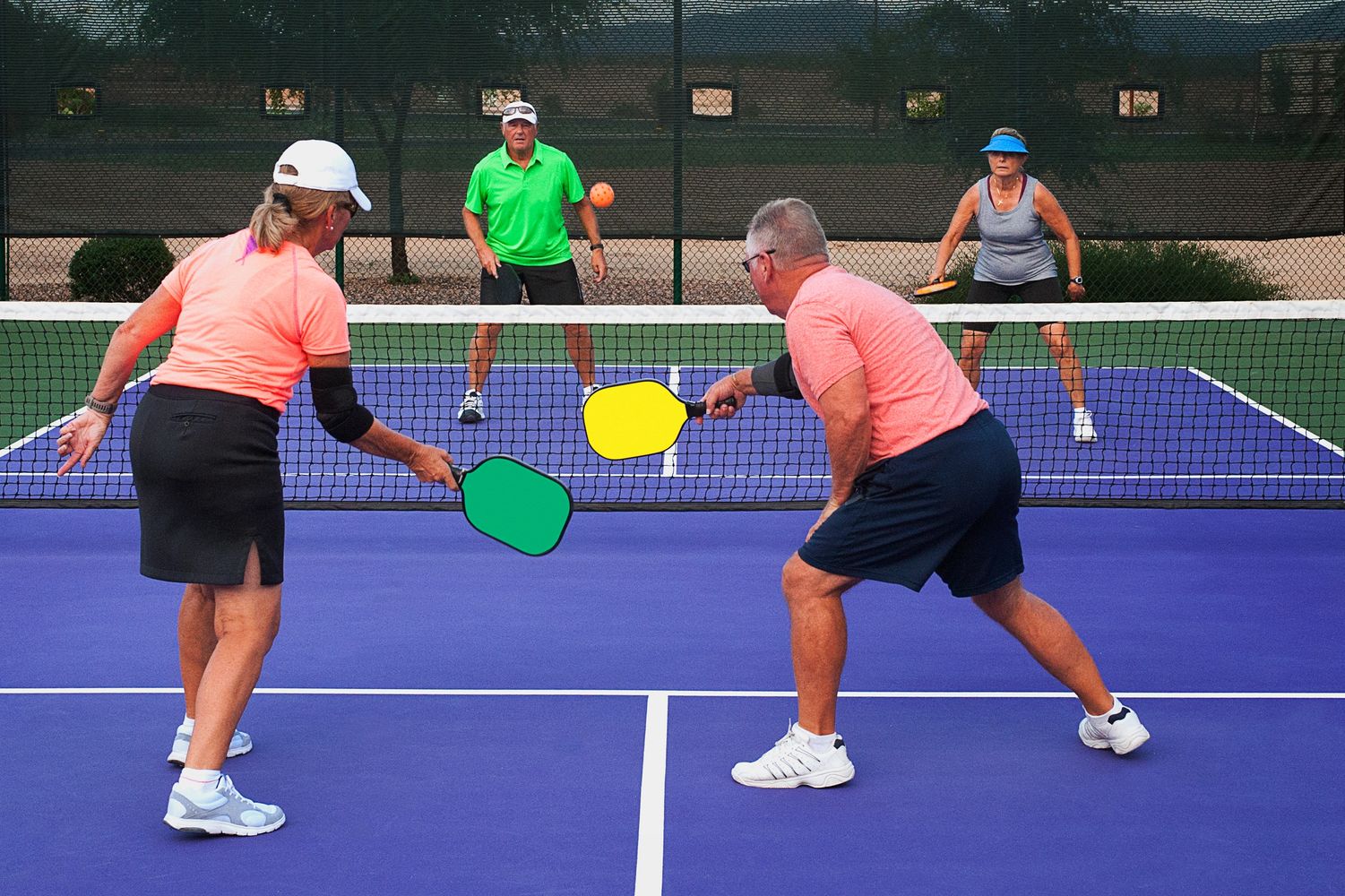 Four athletes playing pickleball, one swings paddle to hit the ball mid-game.

