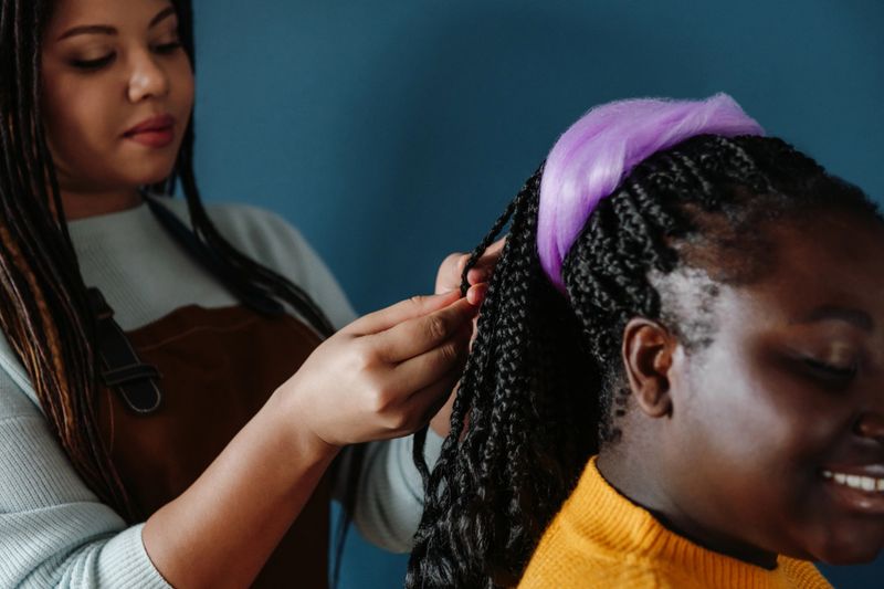 Close-up of confident African hairdresser braiding hair to smiling female customer on blue background