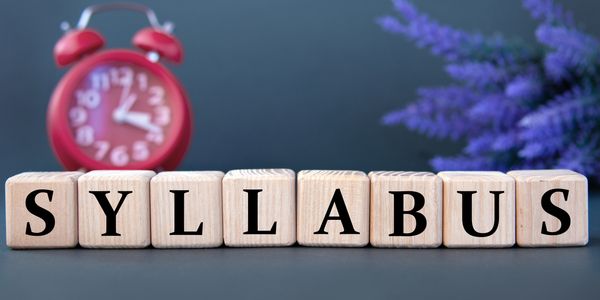 Wooden blocks spell out "SYLLABUS" with an alarm clock and purple flowers in the background.
