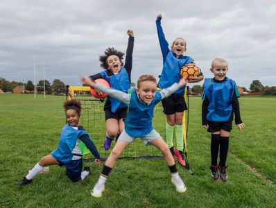 Kids in blue jerseys joyfully jumping on a soccer field with a goal and ball.