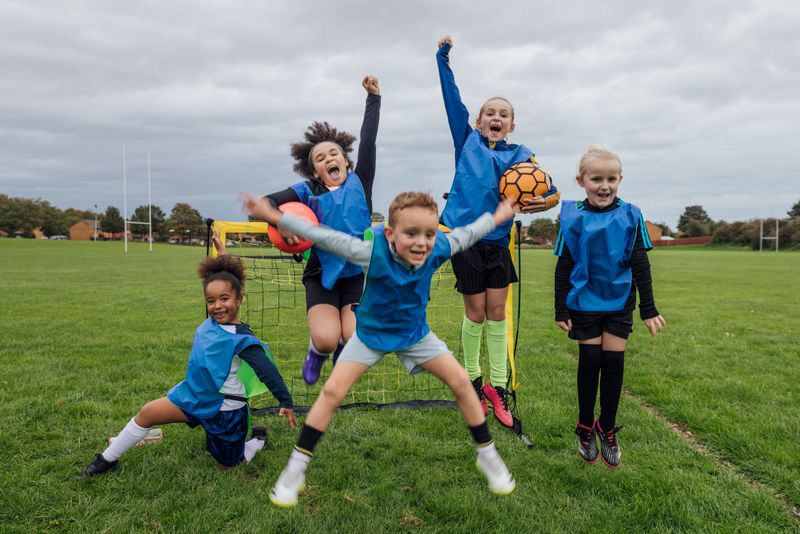 Front view of a small group of boys and girls wearing sports clothing, football boots and a sports bib on a football pitch in the North East of England. They are at football training where they are doing different football training drills. They are jumping up and celebrating with their arms raised in front of a football goal.Videos are available for this scenario.