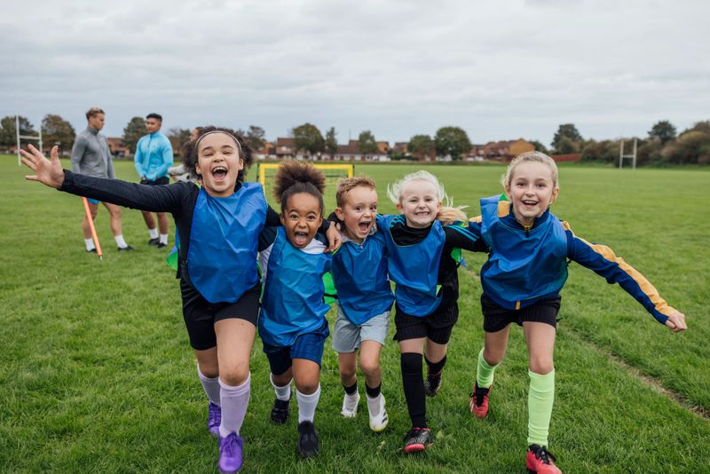 Front view of a small group of boys and girls wearing sports clothing, football boots and a sports bib on a football pitch in the North East of England. They are at football training where they are doing different football training drills. They are running and celebrating with their arms around each other in front of a football goal while smiling and looking at the camera.Videos are available for this scenario.