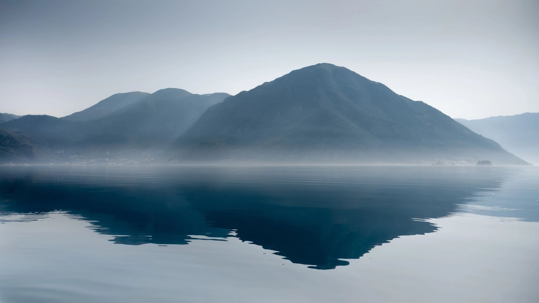 Calm water reflecting misty mountains under a pale sky.
