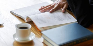 A person reading a book at the table. Next to them is a cup of coffee and two closed books